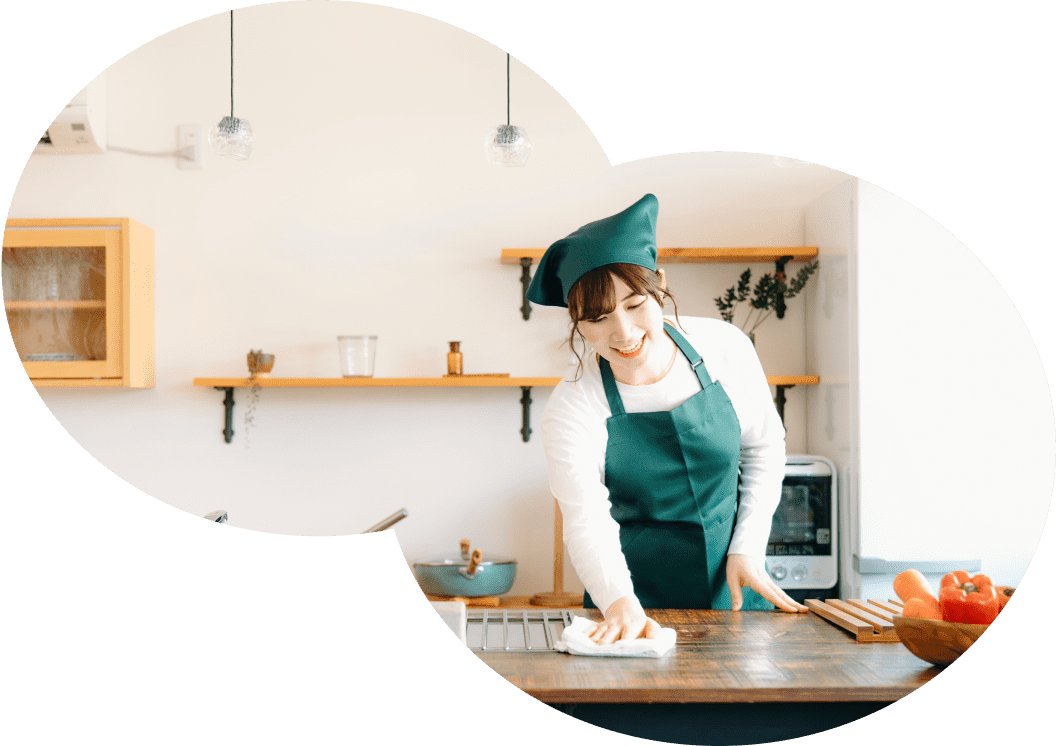 緑色のエプロンと帽子を着用した女性が、明るくモダンなキッチンで木製のカウンターを拭きながら笑顔を見せている様子。背景には棚やキッチン用品が見える