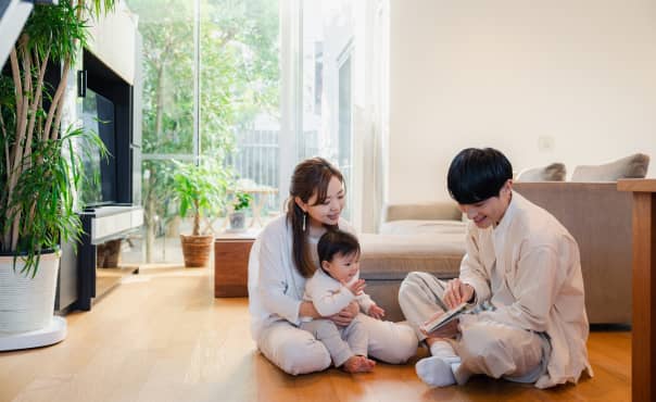 リビングで床に座りながら本を読む若い夫婦と赤ちゃんの家族団らんの様子