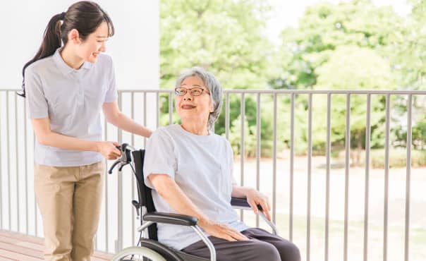 緑豊かな背景をバックに車椅子を押す若い介護士と高齢女性
